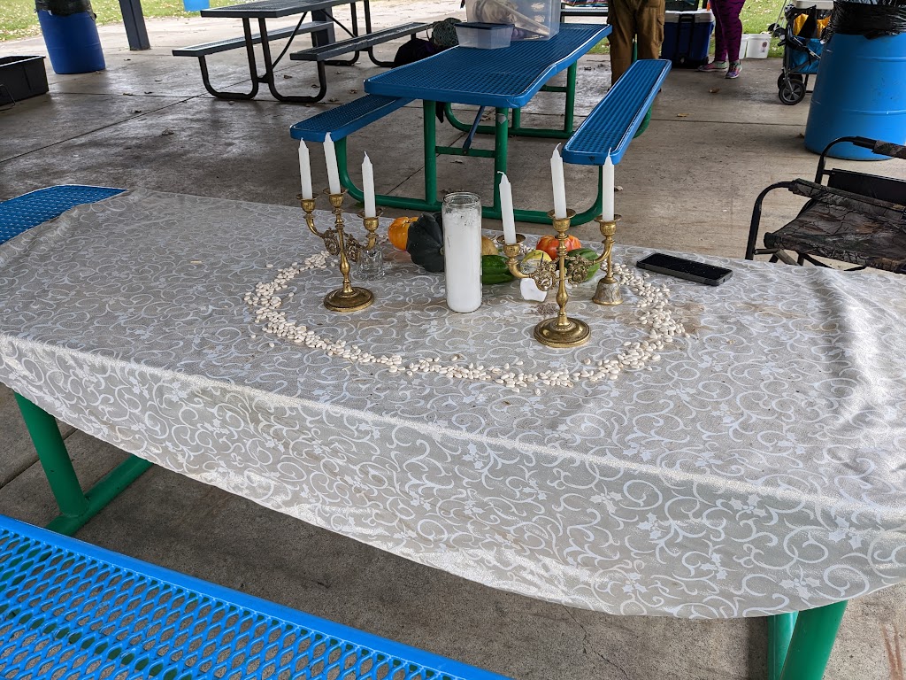 Pagan Pride altar - a swirly beige table cloth over a blue metal picnic table. On the table are 2 candelabras each with three white candles, a white novena candle, small gourds and squashes, and a circle of dried beans